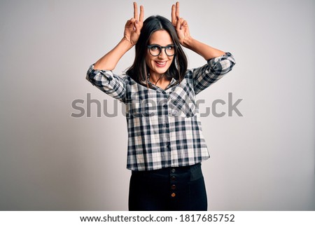 Young brunette woman with blue eyes wearing casual shirt and glasses over white background Posing funny and crazy with fingers on head as bunny ears, smiling cheerful