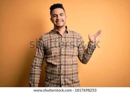Young handsome man wearing casual shirt standing over isolated yellow background smiling cheerful presenting and pointing with palm of hand looking at the camera.