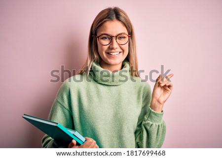 Young beautiful woman wearing glasses holding books over isolated pink background very happy pointing with hand and finger to the side