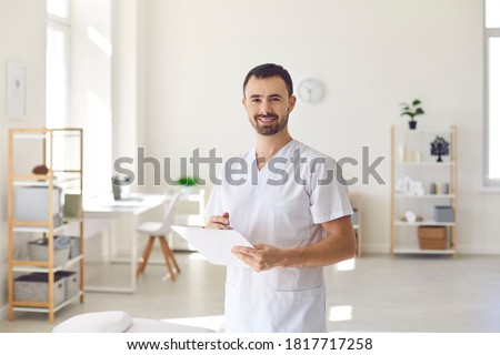 Smiling man doctor chiropractor or osteopath standing with notes and looking at camera with manual therapy clinic interior at background. Professional chiropractor during work concept Royalty-Free Stock Photo #1817717258