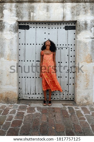 Full length of young African American female in colorful sundress standing against shabby door of old stone building on city street