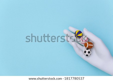 White mannequin hand holding mini balls of different sports on a blue background. Top view