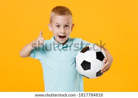 Fun inspired little kid boy 5-6 years old football fan in blue t-shirt cheer up support favorite team, hold in hands soccer ball isolated on yellow background. Sport family leisure lifestyle concept