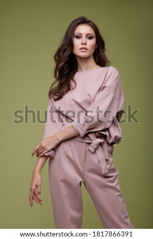 High fashion photo of a beautiful elegant young woman in a pretty pink jumpsuit posing over green background. Slim figure. Studio Shot. Luxurious hair, shiny curls. Femininity and tenderness