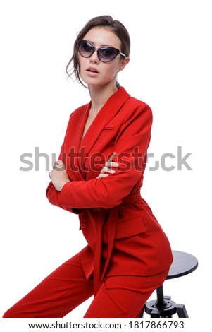 High fashion photo of a beautiful elegant young woman in a pretty red suit, jacket, pants, trousers and sunglasses on white background. Slim figure. Studio Shot. Businesswoman sitting on a black chair