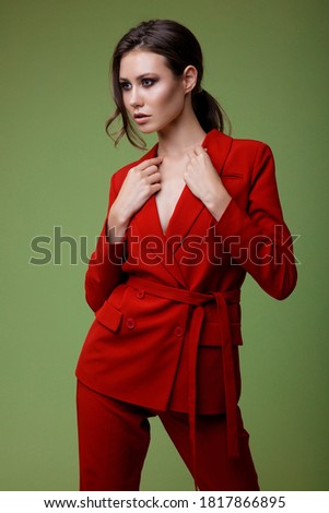 High fashion photo of a beautiful elegant young woman in a pretty red suit, jacket, pants, trousers posing over green background. Slim figure. Studio Shot. Businesswoman. Femininity and success