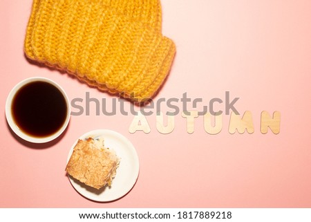 Autumn flat lay with cup of hot coffee, piece of apple pie, warm yellow hat and inscription "autumn" in wooden letters are  on the pastel purple background. 