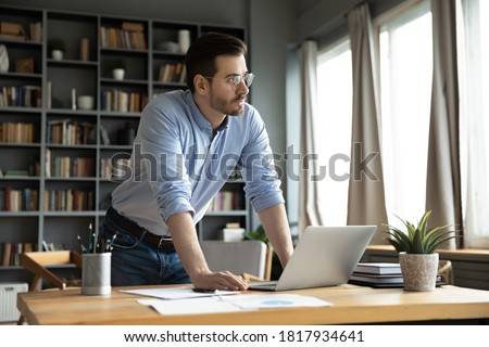 Pensive millennial Caucasian male employee stand at home office desk look in distance think ponder over problem solution. Thoughtful man distracted from computer work make plan decision at workplace. Royalty-Free Stock Photo #1817934641