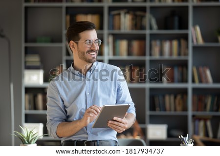 Happy millennial Caucasian man in glasses use tablet look in distance planning or visualizing future success. Smiling young male work on pad gadget, dreaming or thinking. Business vision concept. Royalty-Free Stock Photo #1817934737