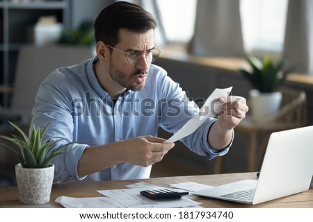 Angry millennial Caucasian guy sit at desk managing financial paperwork frustrated by bill amount. Unhappy young man feel distressed confused paying online, calculating expenses expenditures at home. Royalty-Free Stock Photo #1817934740