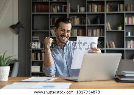 Excited young Caucasian man work on computer feel euphoric reading paper letter correspondence. Happy overjoyed male worker triumph with good pleasant news message in postal paperwork notice. Royalty-Free Stock Photo #1817934878