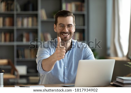 Portrait of smiling young Caucasian man in glasses sit at desk work on laptop recommend online course or training. Happy millennial male show thumb up give recommendation to distant education. Royalty-Free Stock Photo #1817934902