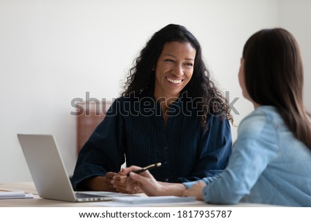 Smiling African American businesswoman talking to colleague, diverse employees brainstorming, sitting at table in office, manager consulting client, using laptop, mentor coach training intern Royalty-Free Stock Photo #1817935787