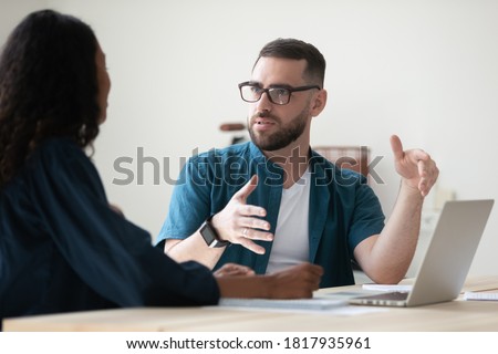 Confident businessman wearing glasses discussing project with colleague at meeting, sitting at table in office, diverse coworkers sharing ideas, working together, mentor coach training intern Royalty-Free Stock Photo #1817935961