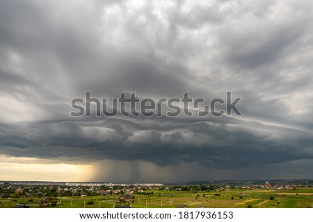 Moody landscape with dark stormy clouds with falling heavy downpour shower rain over distant town buildings in summer.
