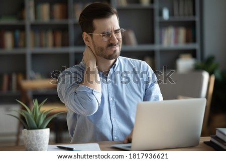 Exhausted young Caucasian male worker sit at desk massage neck suffer from strain spasm muscles. Tired unwell man overwhelmed with computer work sedentary lifestyle struggle with back pain or ache. Royalty-Free Stock Photo #1817936621