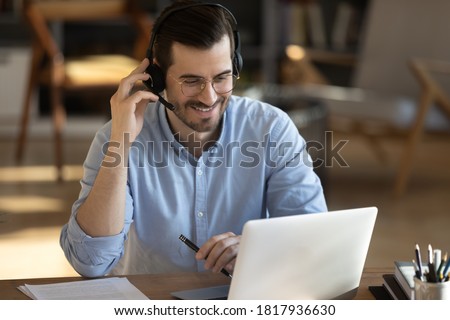 Smiling millennial Caucasian man in headset look at laptop screen work online from home office. Happy young man in headphones watch webinar take course or training on web. Distant education concept. Royalty-Free Stock Photo #1817936630