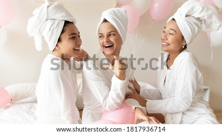 Happy young beautiful caucasian bride preparing for wedding day with excited asian bridesmaids, enjoying fun time after spa procedures together in luxury hotel, wearing towels on heads and bathrobes.