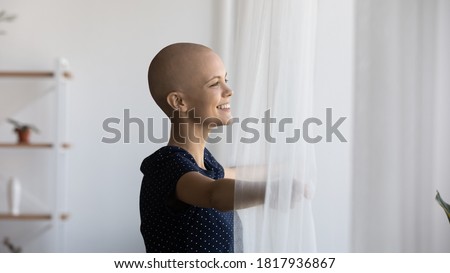Happy young hairless sick woman suffering from oncology open curtains look in window distance dream of recovery. Smiling bald female patient with cancer feel optimistic about remission, good results. Royalty-Free Stock Photo #1817936867