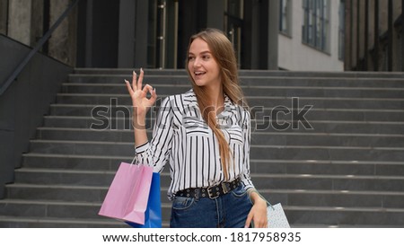 Stylish blonde girl in shirt and jeans holding shopping bags and showing OK sign. Rejoicing good holiday sale discounts. Shopping with low prices, Black Friday sale concept. Mall stairs background