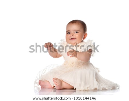 Portrait of sweet infant girl isolated on white in studio. A girl in a beige dress sits barefoot with a cute expressive face and looks at the camera. Place for text.