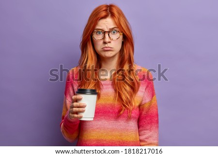 Young blue eyed red haired girl looks with offended sad expression at camera, holds disposable cup of coffee and poses indoor against purple background. Displeased woman dissatisfied by bad weather