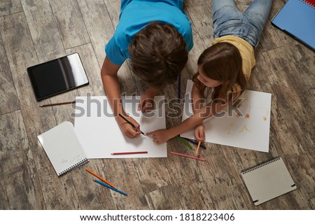 Top view of little caucasian boy and girl lying on the wooden floor at home and drawing on a white sheet of paper with colorful pencils