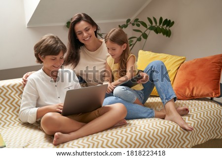 Young beautiful caucasian family, mother and her two cute little kids relaxing on sofa, watching funny videos or cartoons on laptop