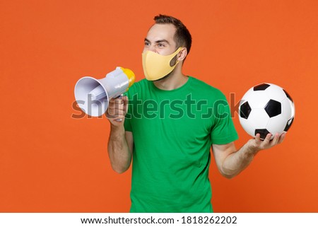 Displeased man football fan in green t-shirt face mask safe from coronavirus support favorite team with soccer ball screaming in megaphone isolated on orange background. People sport leisure concept