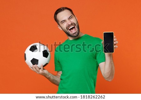Overjoyed young man football fan in green t-shirt cheer up support favorite team with soccer ball hold mobile phone with blank empty screen isolated on orange background. People sport leisure concept