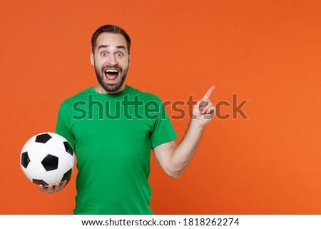 Shocked man football fan in green t-shirt cheer up support favorite team with soccer ball point index finger aside up on mock up copy space isolated on orange background. People sport leisure concept