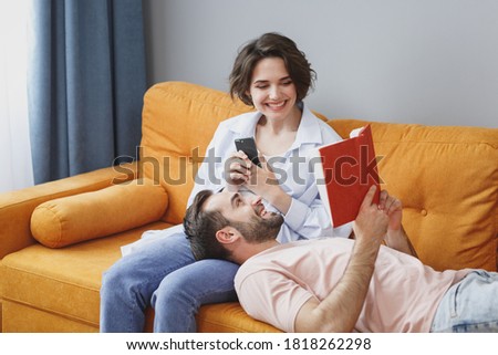 Funny cheerful smiling young couple two friends man woman 20s wearing casual clothes sitting lying on couch using mobile cell phone reading book resting relaxing spending time in living room at home