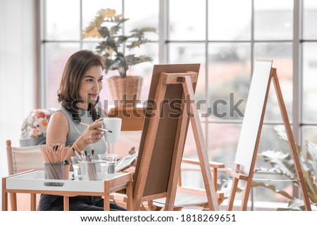 Beautiful lady Asian woman in casual clothing holding coffee mug sitting and use a brush to pain picture in the room. Idea for hobby, relaxation or artist work from home. 