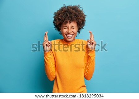 Curly haired positive Afro American woman closes eyes and crosses fingers hopes for better wears orange jumper poses against blue background smiles broadly. Agitated female makes wish poses indoor