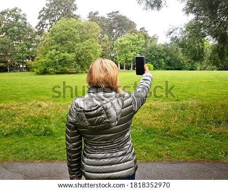 Latin woman in a gray anorak making a selfie with the mobile phone in the middle of a landscaped path take from behind