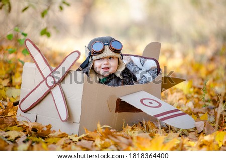 A happy child dreams of traveling in an airplane and plays an airplane pilot. The kid sits in a cardboard plane and pretends to be a pilot in the park in the fall.