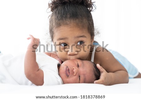 Cute African American little girl kissing on newborn baby cheek on white bed at home. Little girl takes care of infant baby with kindly