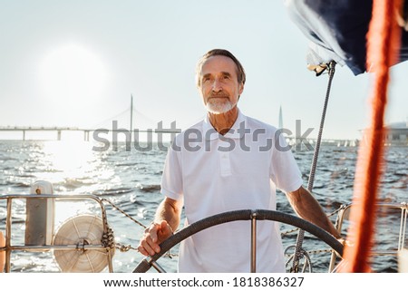 Mature man steering a private yacht while on vacation. Bearded yachtsman standing on his sailing boat looking at distance. Royalty-Free Stock Photo #1818386327