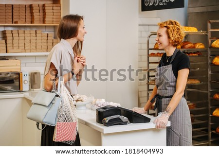 Pretty young female baker standing behing cash register talking to client, taking her order, charmingly smiling. Side view.