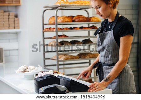 Pretty young female baker standing behing cash register calculating overal cost of the order. Side view. Cropped, half head.