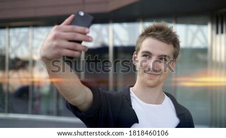 Young blond guy making selfie on background of glass building, casual guy with phone lens flare. Portrait of smiling guy making selfie on background of business building