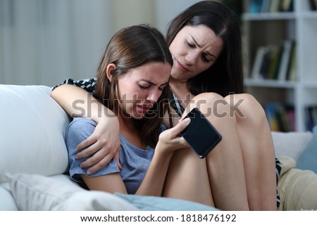 Sad teen with phone being comforted by her sister on a couch in the living room at home  Royalty-Free Stock Photo #1818476192