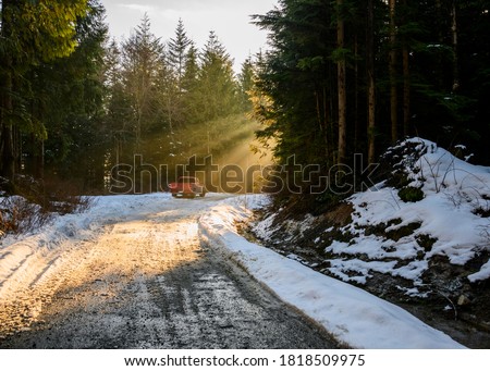 Beautiful nature woodland scene. Springtime foliage. sun behind the forest. Sun rays hitting the trees, beaming down. Snow on the forest road. Dark tree silhouettes. Red pick up truck. Royalty-Free Stock Photo #1818509975