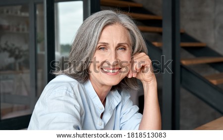 Smiling stylish mature middle aged woman sitting at workplace, portrait. Happy older senior businesswoman, 60s grey-haired lady executive manager looking at camera sitting at office table. Headshot.