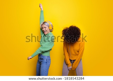 Cute women with headphones dancing on a yellow studio wall full of emotions wearing warm sweaters and jeans