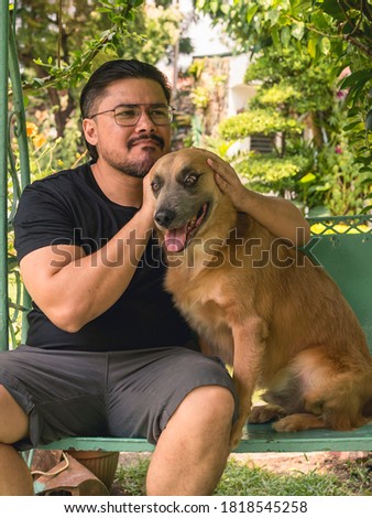 A man covers his pet dog's ears while both are sitting on an outdoor patio swing. Best friends concept.