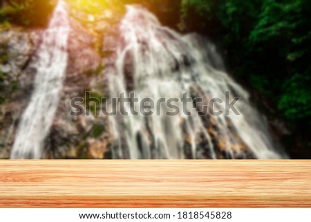 Blurred beautiful steep rocky waterfall with flowing water in forest that behind top of empty wooden table. Scenic landscape and tabletop with empty space for montage product display at seaside.