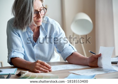 Mature old adult elegant woman fashion designer drawing creative sketches on table. Beautiful sophisticated middle aged grey-haired lady entrepreneur creating new fashion design cloth in atelier.