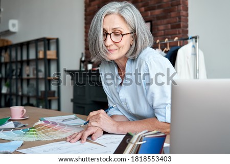 Middle aged stylish woman fashion designer drawing sketches in studio office. Mature old adult elegant grey-haired lady dressmaker small business owner creating new fashion design cloth in atelier.