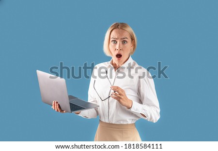 Portrait of shocked businesswoman holding laptop and looking at camera over blue studio background. Female office employee surprised over unexpected news or gossip, astonished at career achievements Royalty-Free Stock Photo #1818584111